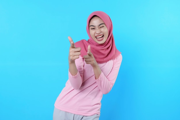 Smiling asian woman with her finger pointing isolated on light blue background wearing hijab with pink t shirt