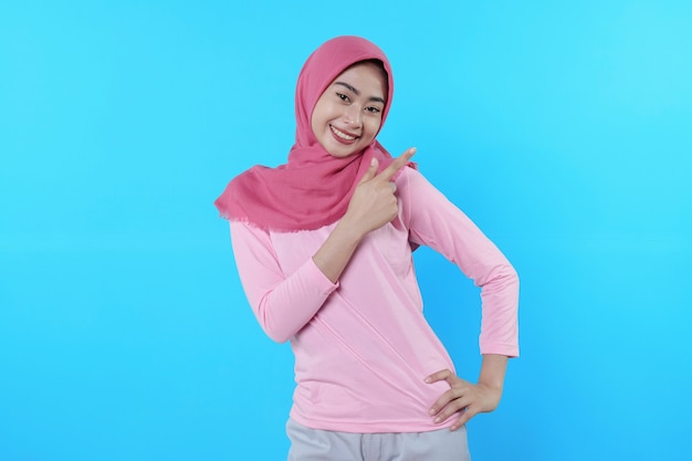 Smiling asian woman with her finger pointing isolated on light blue background wearing hijab with pink t shirt