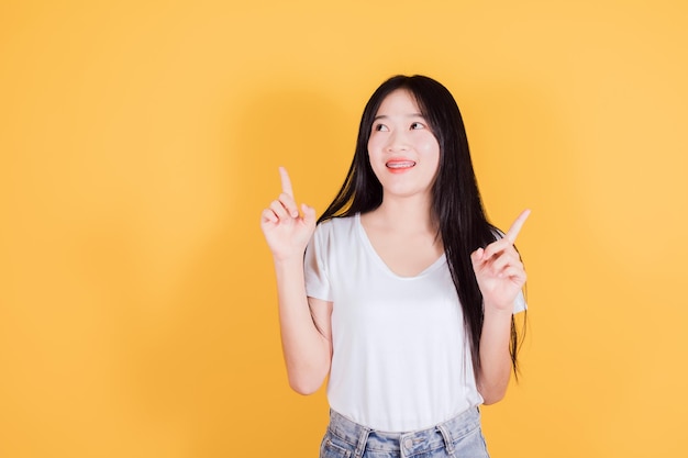 Smiling Asian woman with long dark hair wears white tshirt pointing finger to the side on yellow background