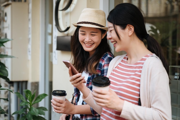 Smiling asian women tourist show to laughing friends funny news photo at mobile phone having fun together while relax lean on windowsill outdoor with coffee cup. two girls joy look cellphone screen