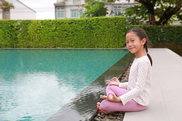 Smiling Asian young girl child practicing mindfulness meditation sitting near the pool with looking camera