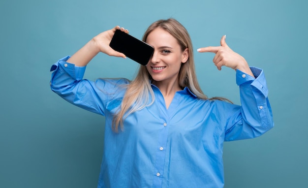 Smiling attractive blond girl showing blank smartphone screen with mockup for web page on blue