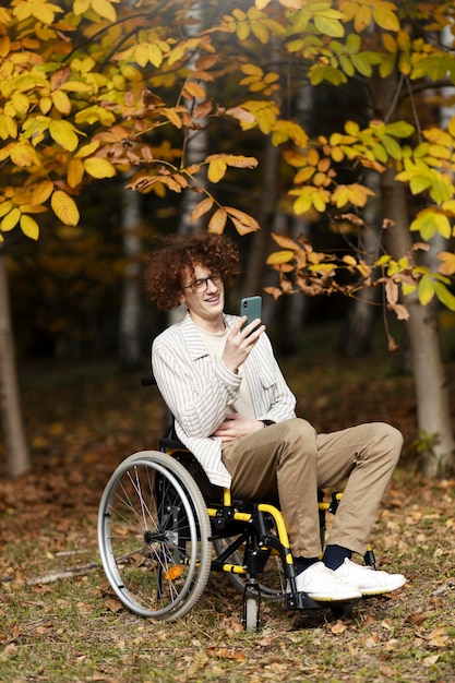 Smiling attractive guy in glasses sits in a wheelchair and holds a smartphone in his hand A disabled boy outdoors under a tree with yellow leaves