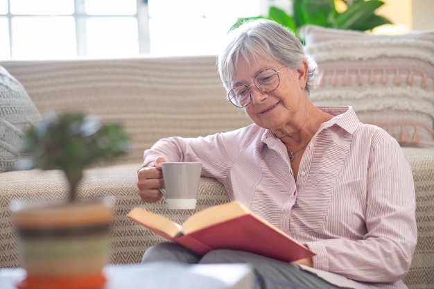 Smiling attractive senior woman relaxing sitting on the floor reading a book holding a cup of coffee or tea Caucasian mature lady studying learning at home