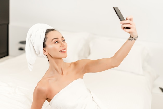 Smiling attractive woman doing selfie on the bed in bedroom after shower 