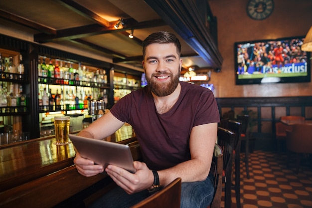 Smiling attractive young man using tablet and drinking beer in pub