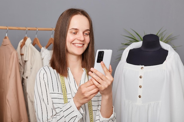 Smiling attractive young professional dressmaker using smartphone while working in atelier searching new ideas in internet browsing networks fashion blog sewing dresses for new collection