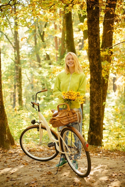 Smiling attractive young woman in autumn park holding flowers standing near her retro bike Blonde en