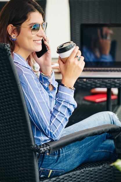 Smiling attractive young woman in sunglasses sitting at table in outdoor cafe, drinking coffee and talking on phone