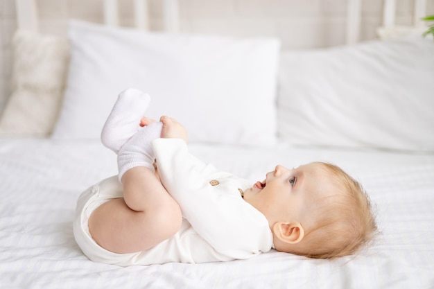 Smiling baby 6 months old blond boy lies on a large bed in a bright bedroom and plays with his legs in a cotton bodysuit the concept of children's goods