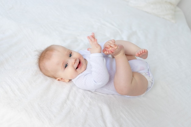 Smiling baby girl six months old plays with her legs lying on her back on a white cotton bed in the bedroom of the house and laughs.