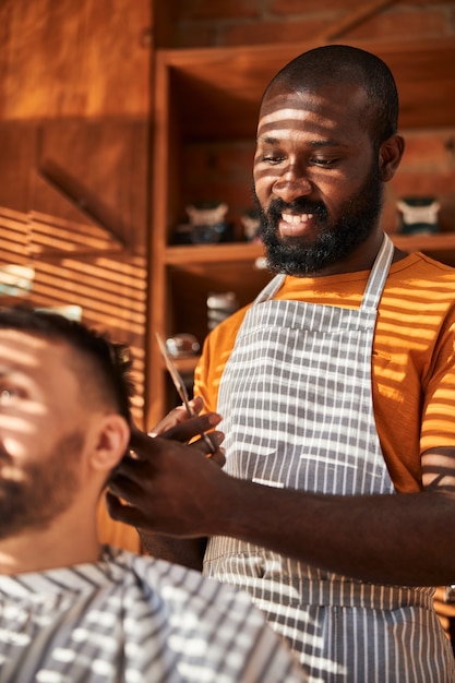 Smiling barber making haircut for client in barbershop