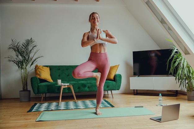 Photo smiling barefoot woman in sportswear balances on one leg on yoga mat in living room