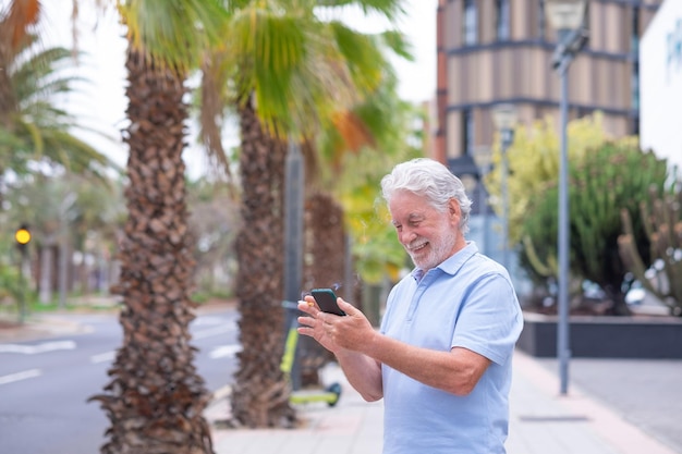 Smiling bearded caucasian senior man using mobile phone standing outdoor in the city street smoking a cigarette Palm trees on background