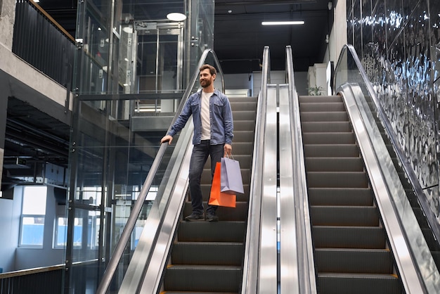 Smiling bearded man carrying shopping bags while going down on escalator of shopping mall