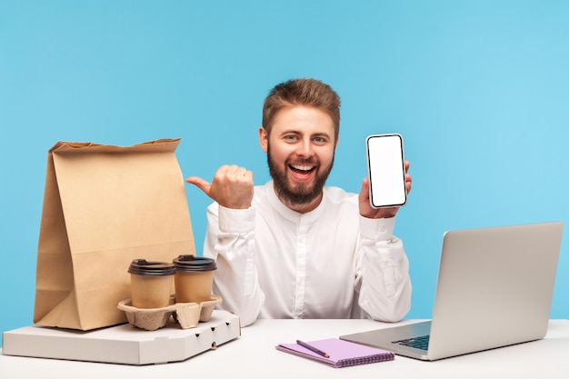 Smiling bearded man holding white screen smartphone pointing finger at order with fast food on his workplace desk, online order fast delivery. Indoor studio shot isolated on blue background