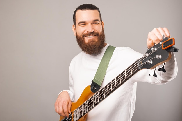 Smiling bearded man is holding a wooden colour bass guitar in a studio