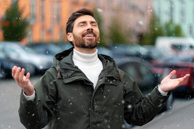 Smiling bearded man with closed eyes stretched out his hands and sincerely rejoices Man outdoors with snow falling on him