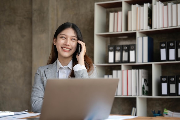 Smiling beautiful Asian businesswoman analyzing chart and graph showing changes on the market and holding smartphone at office