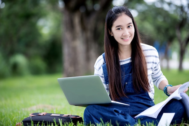 Smiling beautiful Asian girl reading book and working at tree on park in summer for relax time