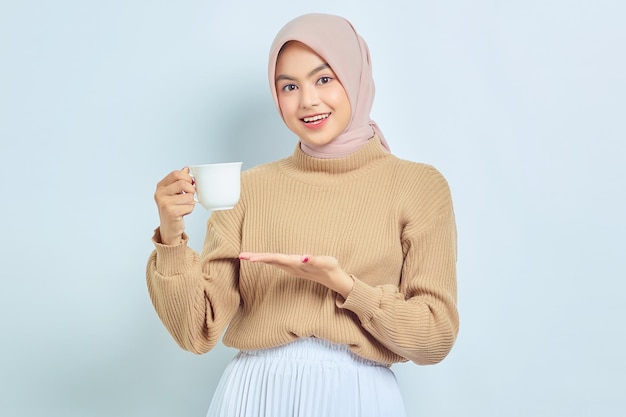 Smiling beautiful Asian muslim woman in brown sweater showing mug of coffee cup in hands isolated over white background People religious lifestyle concept