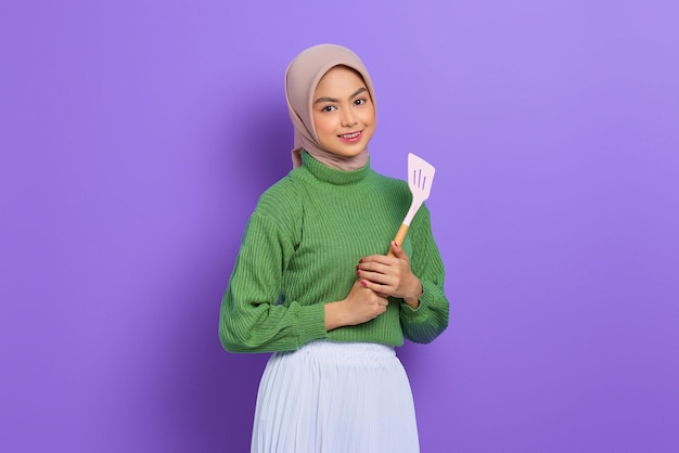 Smiling beautiful Asian woman in green sweater and hijab holding spatula isolated over purple background