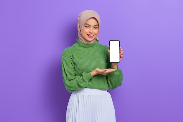 Smiling beautiful Asian woman in green sweater and hijab showing blank screen mobile phone isolated over purple background