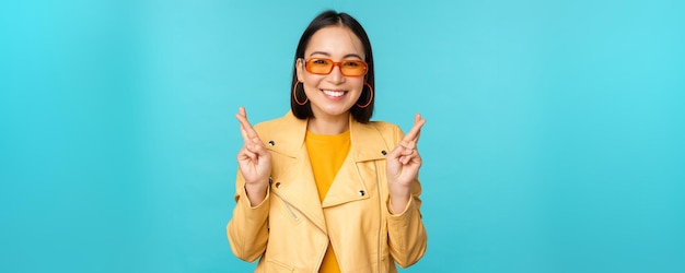 Smiling beautiful asian woman wishing cross fingers for good luck and looking hopeful standing over blue background Copy space