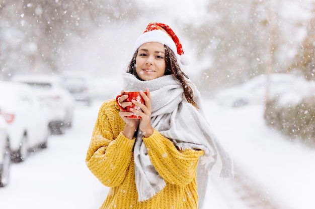 Smiling beautiful girl in a Santa hat stands outside in the snow. Merry Christmas background