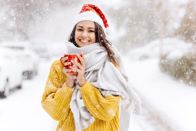 Smiling beautiful girl in a Santa hat stands outside in the snow. Merry Christmas background