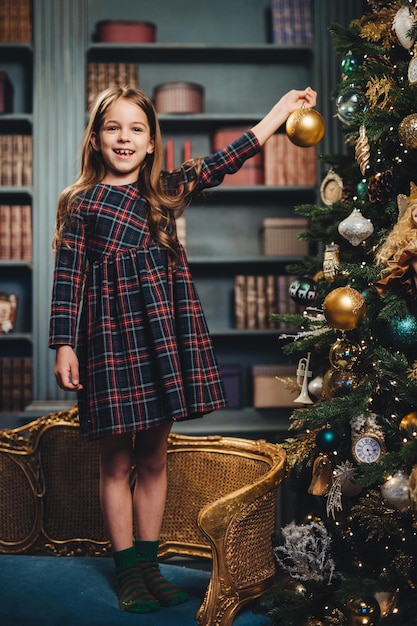 Smiling beautiful little girl stands on armchair holds glass ball decorates New Year tree being happy to do it Adorable small child busy with decoration of Christmas tree Season and celebration