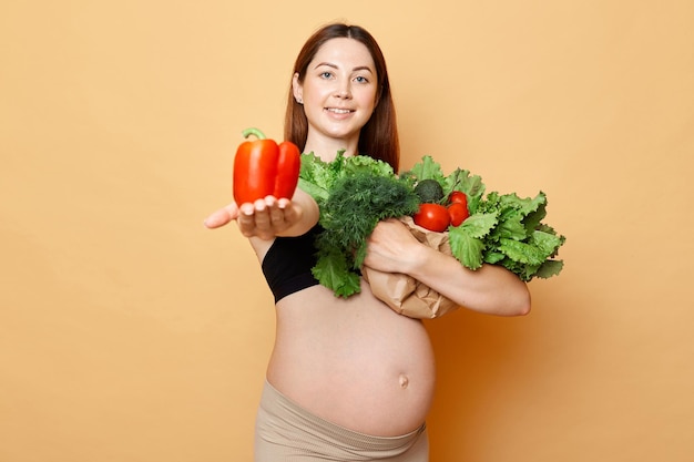 Smiling beautiful pregnant woman embraces fresh vegetables posing isolated over beige background future mother with bare belly showing offering organic pepper