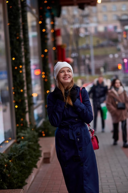 Smiling beautiful woman in the evening street near decorated showcase Stylish winter casual clothes City lights Festive city