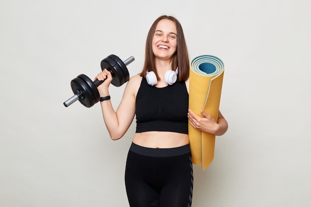 Smiling beautiful woman holding rolled fitness mat and barbel dressed in activewear isolated over gray background looking at camera with smile enjoying workout