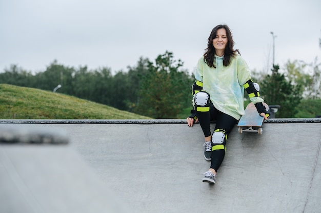 Smiling beautiful woman skater resting on a deck. She wears some protective gear. Frontal view. Nature in background.