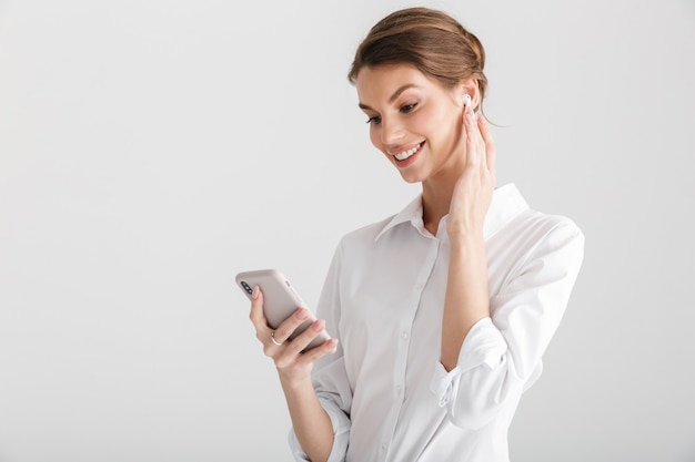 smiling beautiful woman using wireless earphone and mobile phone isolated over white background