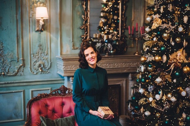 Smiling beautiful woman wears nice dress, sits near Christmas tree in room, holds present