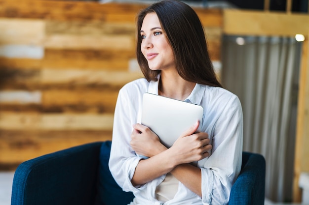 Smiling beautiful woman in white shirt sits on an armchair and squeezes smart tablet tightly.