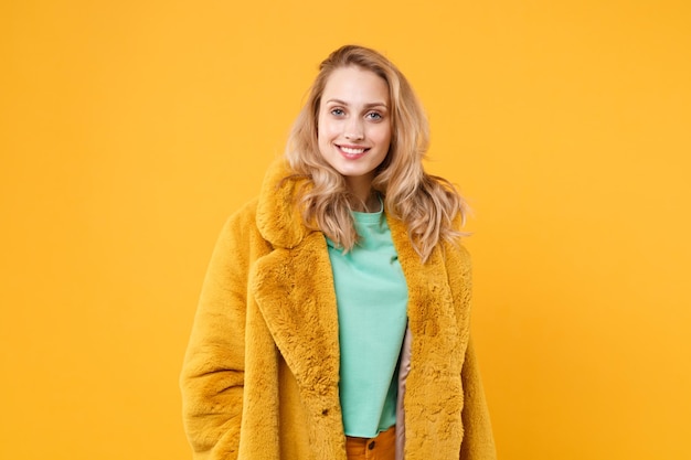 Smiling beautiful young blonde woman girl in yellow fur coat posing isolated on orange wall background studio portrait. People sincere emotions lifestyle concept. Mock up copy space. Looking camera.