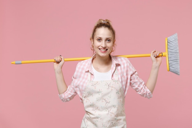 Smiling beautiful young woman housewife in casual clothes, apron doing housework isolated on pastel pink wall background studio portrait. Housekeeping concept. Mock up copy space. Hold in hands broom.