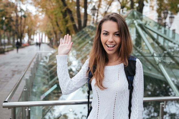 Smiling beautiful young woman waving and saying hello to you in park