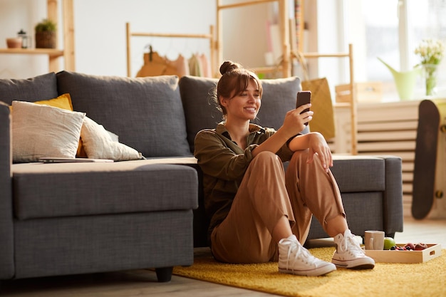 Smiling beautiful young woman with hair bun sitting on carpet and reading post on phone while having