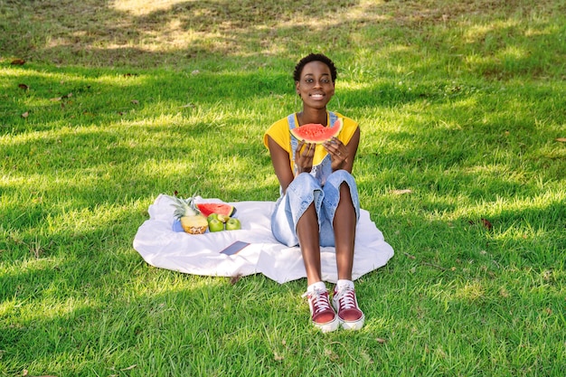 Smiling black African woman sitting in an outdoor park while eating a watermelon and more fruit