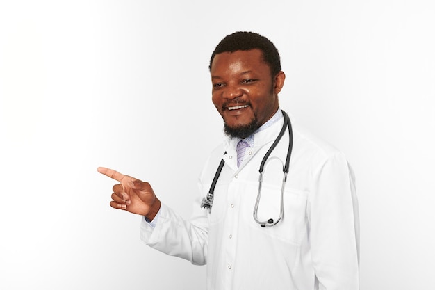 Smiling black bearded doctor man in white coat with stethoscope points finger white background