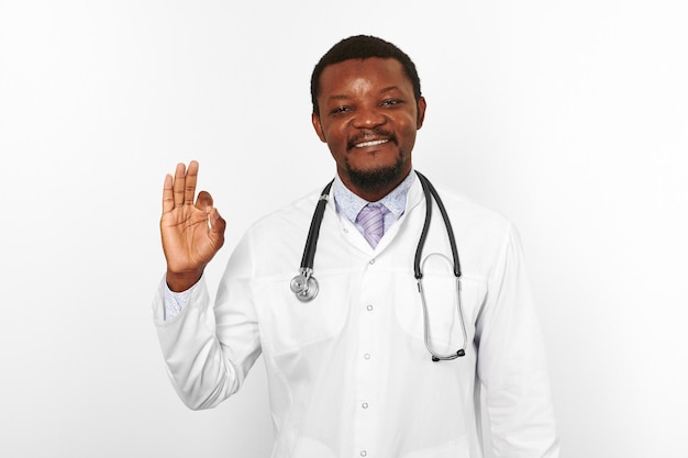 Smiling black bearded doctor man in white robe with stethoscope shows OK gesture white background