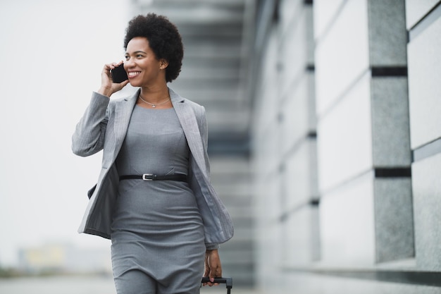 A smiling black business woman talking on her smart phone while walking down a street, in front a corporate building.