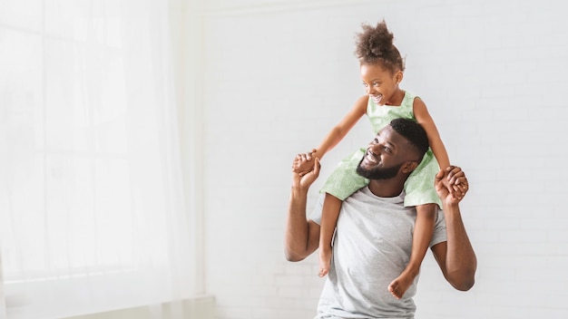 A smiling black father gives his daughter a piggyback ride in a bright white room the little girl is