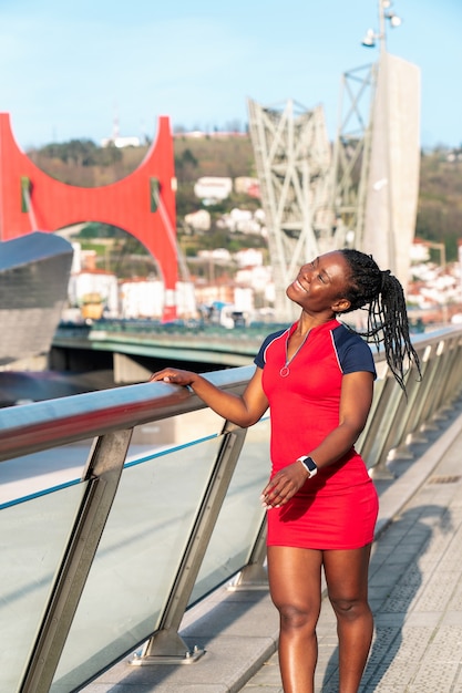 Smiling black girl of afro ethnicity walking down the city street in a colourful red sports dress with her hair in plaits