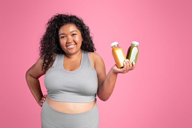 Smiling black obese lady in sportswear show bottles with juice or smoothie drink recommendation for