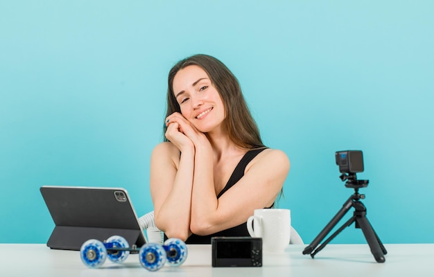 Smiling blogger girl is looking at camera by holding hands together on cheek on blue background